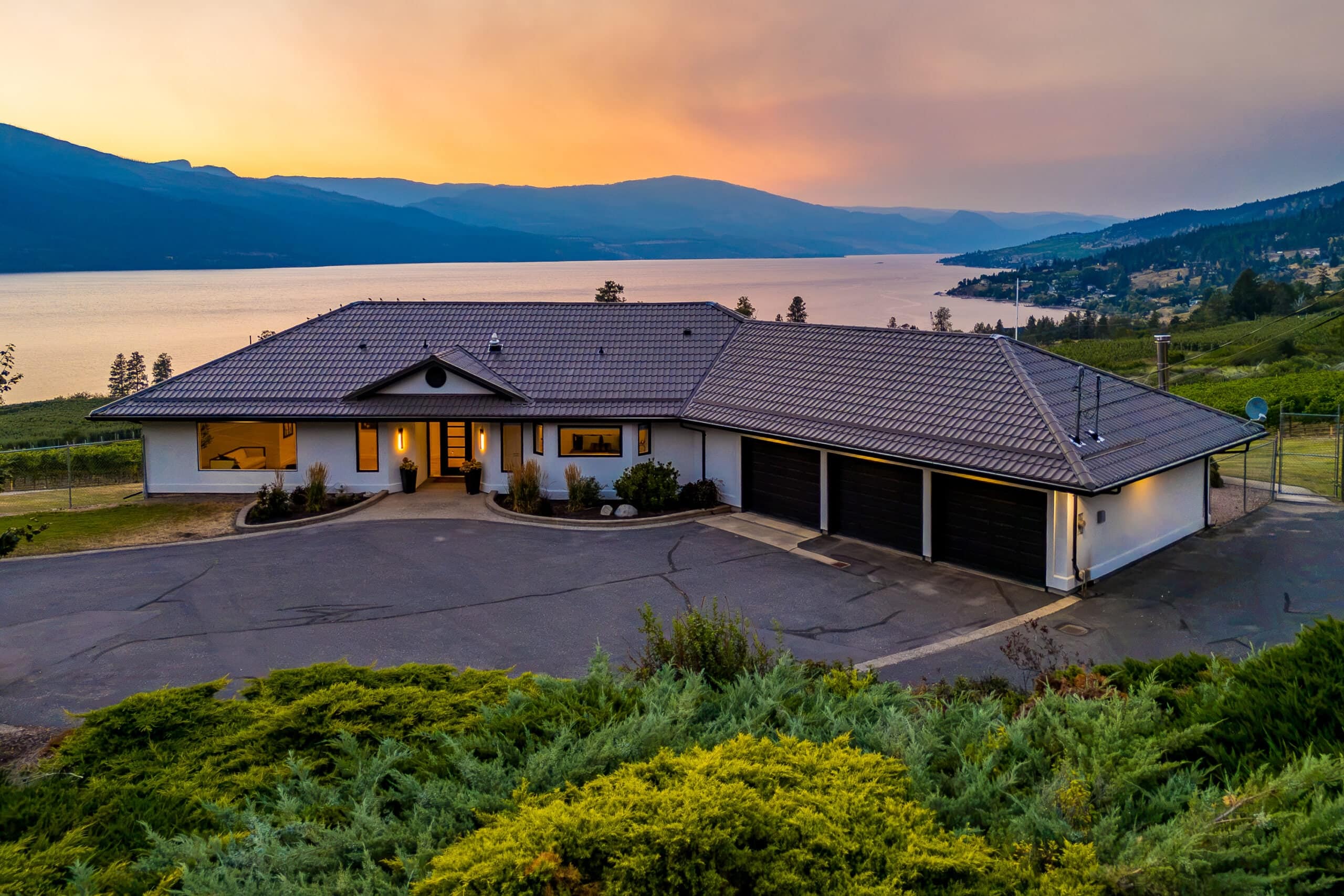 1090 Camp Road at sunset: a lit house above its gardens, Okanagan Lake and the hills beyond