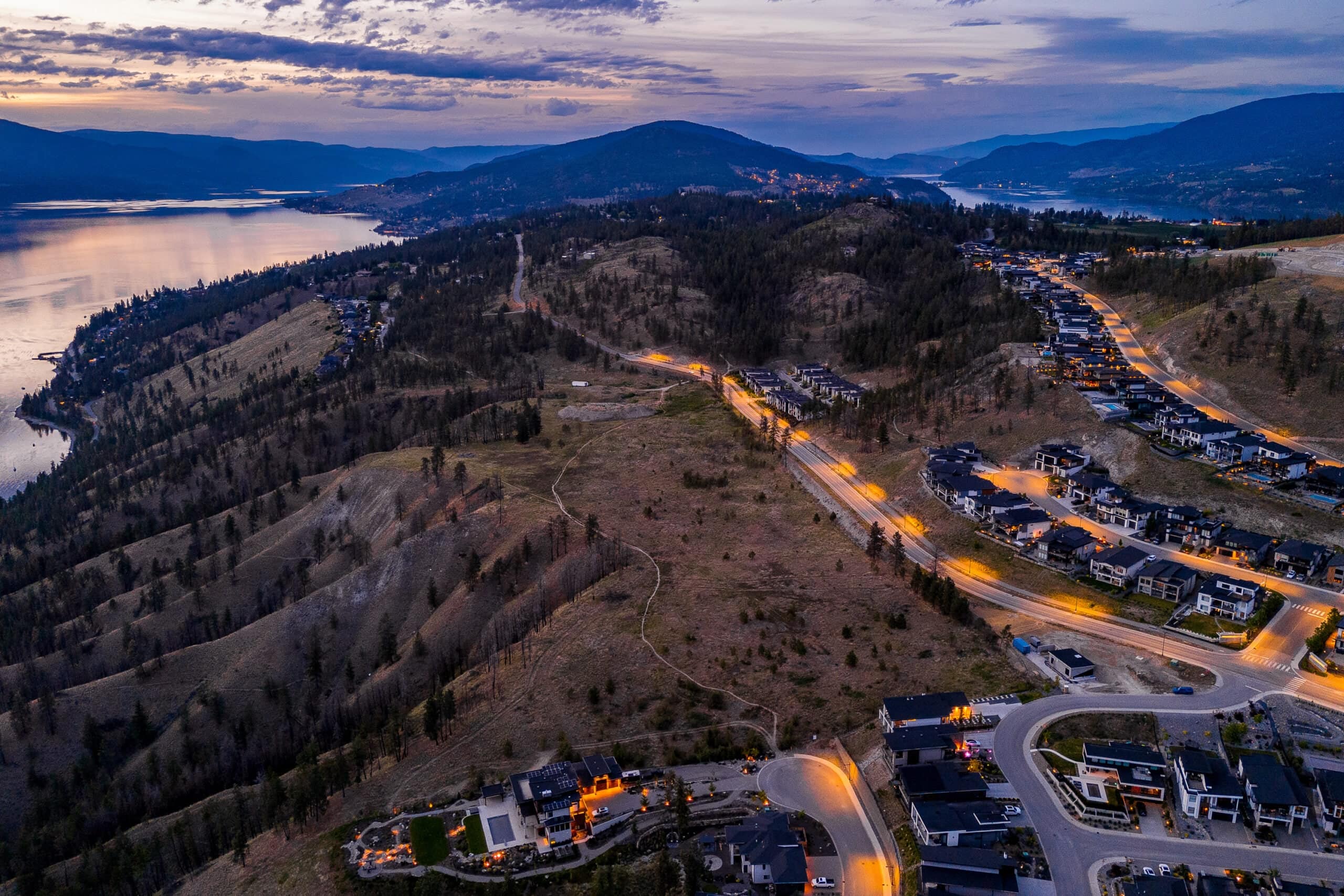 Lakestone's lit roads at dusk winding past the dark slope of Tyndall Bench, Okanagan Lake at the left